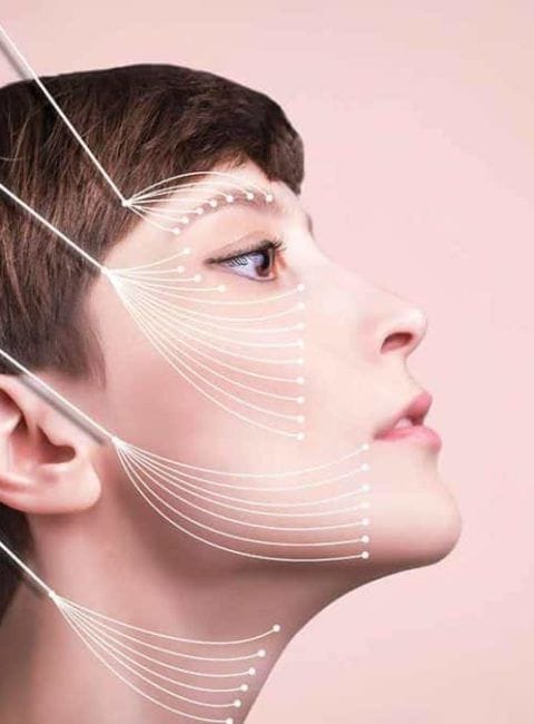 A profile view of a woman's face against a soft, solid pastel pink background, showcasing professional aesthetic treatment mapping. The lighting is bright, even, and clinical, highlighting smooth, clean skin with very subtle, natural makeup focusing on the eye area. The subject displays feminine facial features with a well-defined jawline, nose, and brow structure. The anatomy of the face is emphasized by white graphic lines radiating from multiple points toward the brow, cheeks, jawline, and neck, illustrating threads or aesthetic placement. The subject is wearing no visible clothing, focusing entirely on the head and neck profile.