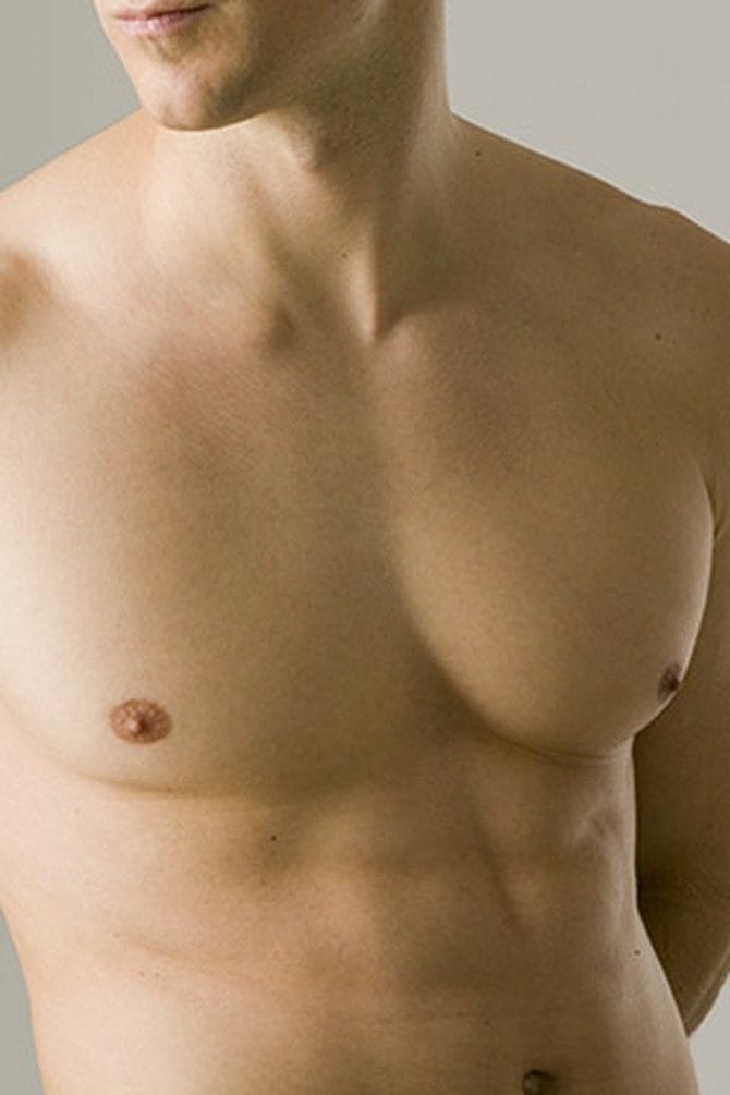 A close-up studio shot of a shirtless man's torso. The lighting is soft and even, highlighting well-defined pectoral muscles and a toned abdomen. The subject exhibits masculine anatomy, with no visible makeup, against a clean, neutral, light grey background. He is not wearing any clothing.