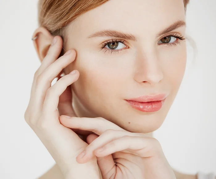 A close-up portrait of a woman with feminine facial features, featuring soft, natural lighting that highlights her radiant complexion. Her makeup is minimalist and natural, emphasizing her clear skin, long lashes, and soft pink lips. She has a delicate facial anatomy with high cheekbones, and her hands are gently cradling her face. The background is a clean, bright, and solid white, focusing entirely on her face and expressions without distractions; no specific clothing details are visible in this head-and-shoulder frame.
