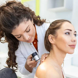 A female dermatologist with dark curly hair, wearing a white lab coat over a rust-colored top, uses a dermatoscope to examine the shoulder of a female patient with light brown hair. The lighting is soft and professional, typical of a clinical setting. The doctor has natural-looking makeup, and both women exhibit feminine features. The background is a clean, blurred medical office with light colors, focusing the attention on the skin examination, anatomy, and the doctor's precise posture.