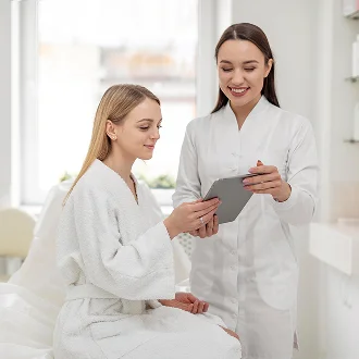 A bright, professional medical aesthetic clinic setting with soft, natural lighting. A female patient with natural makeup, wearing a plush white bathrobe, sits comfortably while a smiling female practitioner in a crisp white medical uniform stands beside her, explaining treatment plans using a tablet. Both figures are feminine, with relaxed postures. The background is a clean, modern, out-of-focus clinic interior with neutral tones. The composition highlights a friendly, expert-patient consultation.