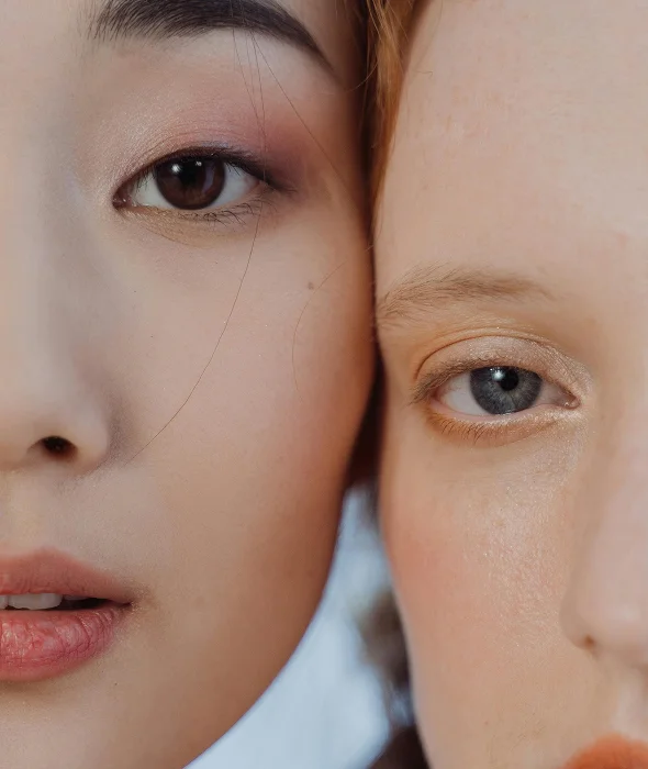 Close-up beauty portrait of two feminine faces side-by-side: a woman with East Asian features and dark brown eyes on the left, and a woman with fair skin, light hair, and grey-blue eyes on the right. Both feature soft, natural, glowing makeup with subtle earth-toned eyeshadows. The lighting is soft, diffused, and even, highlighting skin textures. The background is a clean, bright, and out-of-focus neutral tone. No clothing is visible. The composition focuses exclusively on the facial anatomy, emphasizing the contrast in skin tones and eye colors between the two subjects.