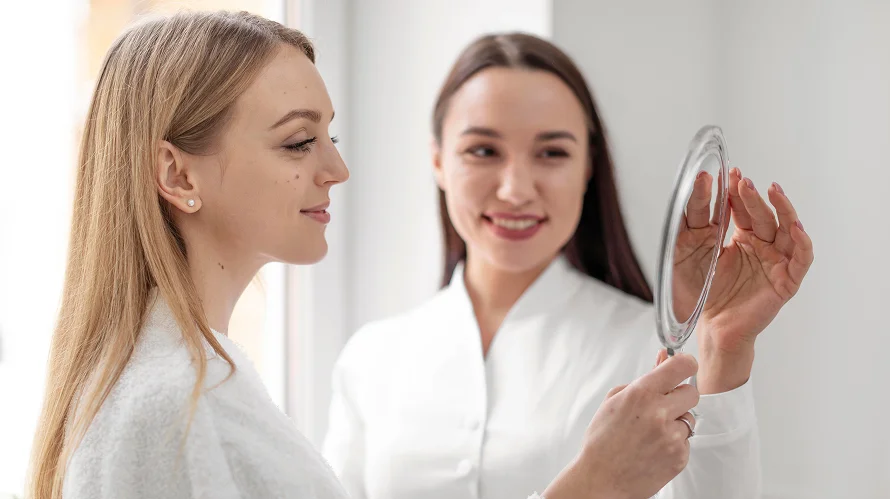 A female doctor with long dark hair, wearing a professional white medical coat, holds a round hand mirror for a fair-skinned female patient with blonde hair and a white bathrobe to see her face. Both women have feminine features, natural-looking makeup, and soft skin textures. They are in a well-lit, clinical environment with a bright, clean, white background that enhances the soft, professional lighting of the scene. The patient is shown in profile while the doctor is slightly out of focus in the background, both radiating a calm and professional demeanor.