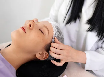 A side-profile view of a female patient with natural skin lying down during a skincare treatment, wearing a disposable hair net. A professional with dark hair, dressed in a white medical coat, is carefully adjusting the hair net. The lighting is soft and bright, creating a clean, clinical aesthetic. The background is a simple, neutral-toned wall, emphasizing the focus on the facial anatomy and professional care.