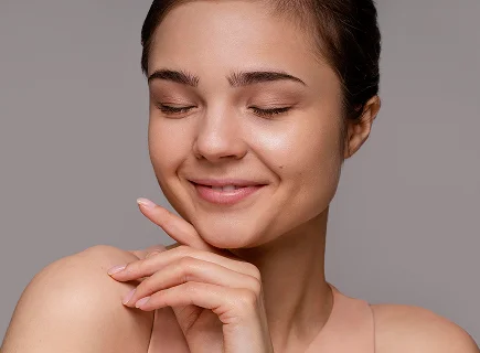 A close-up portrait of a young feminine-presenting individual with smooth skin and a serene expression. Their eyes are gently closed, and their face is subtly illuminated with soft, diffused lighting that enhances their facial features. The makeup is natural and minimal, emphasizing a fresh and glowing complexion with a slight highlight on the cheekbones. The background is plain and neutral, likely a soft gray or light pastel shade, ensuring the focus remains on the subject. The individual is wearing a simple, strapless top, allowing visibility of their collarbone and shoulders, which adds to the elegant and calm aesthetic of the image.