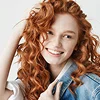 A smiling young woman with voluminous, vibrant curly red hair radiating feminine energy, captured in bright, soft lighting against a clean white background. She is wearing a casual blue denim jacket over a white top, with natural-looking makeup that accentuates her gentle facial features and joyful expression. Her hand is gracefully touching her hair, highlighting the natural texture of her curls.