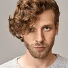 A headshot of a masculine man with wavy, light brown hair styled to the side, soft stubble on his face, and a natural, minimal makeup look. The lighting is soft and even, highlighting his facial anatomy, set against a neutral, plain light gray background. He is wearing a simple, textured light-colored knitted sweater.