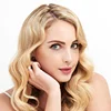 A portrait of a woman with a highly feminine appearance, featuring long wavy blonde hair, soft natural makeup with emphasis on her eyes and lips. She is captured with studio lighting against a clean white background. Her anatomy shows delicate facial features, and she is wearing a simple white garment with thin straps, showcasing a balanced and elegant composition.