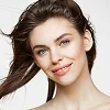 Close-up portrait of a woman with a feminine facial structure, featuring soft, radiant natural lighting. She has a natural makeup look with defined brows, subtle eye makeup, and a soft pink lip. Her hair is slightly tousled by a gentle breeze. She exhibits soft, youthful anatomical features with high cheekbones. The background is a clean, neutral white, and the subject is wearing a simple, understated top, focusing the viewer's attention on her expressive face.