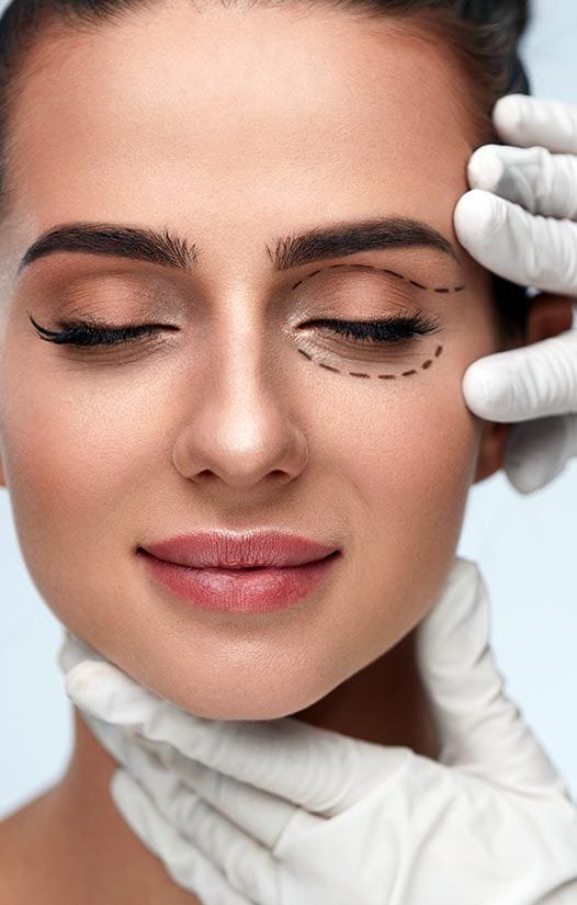 Close-up portrait of a woman with feminine features and natural makeup, including soft eyeshadow and pink lipstick. A plastic surgeon's hands, wearing white medical gloves, are touching her face. Dashed surgical guide lines are drawn around her eye area for a blepharoplasty procedure. The lighting is bright and clinical, highlighting the smooth skin texture and facial anatomy. The background is a clean, solid light blue, and no clothing is visible in the frame.