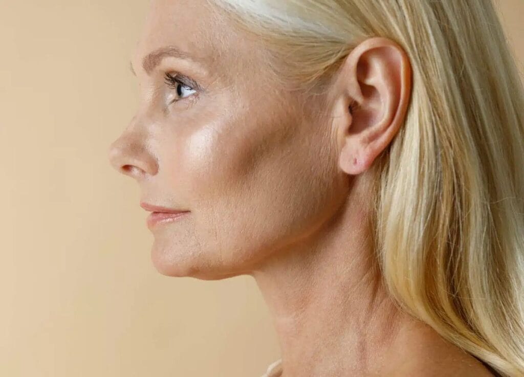 A close-up side profile of an elegant, mature woman with blonde hair, soft natural lighting highlighting her facial features, wearing minimal, fresh-faced makeup that emphasizes a feminine aesthetic, set against a smooth, neutral beige background; her anatomy shows graceful, aged skin texture and sharp bone structure, dressed in a simple, light-colored garment.
