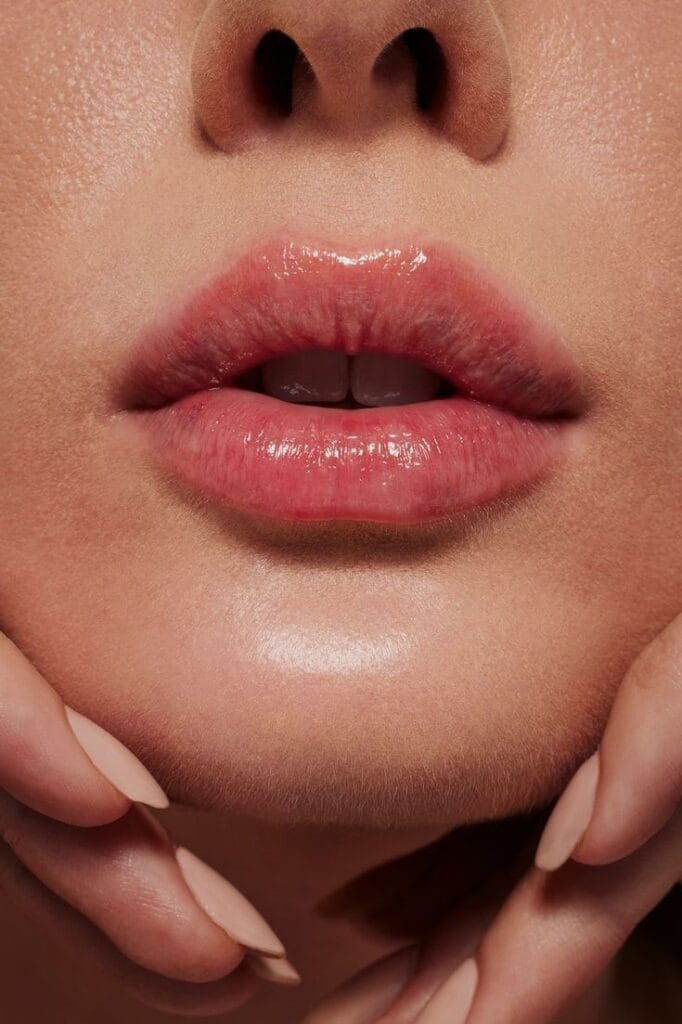 Close-up shot of feminine lips with a glossy, natural-toned finish. The lighting is soft and even, highlighting the skin's texture and a slight shimmer on the chin. The makeup is minimalist and natural, emphasizing the lips' hydrated appearance. The subject is holding their chin with hands featuring nude-colored, manicured almond-shaped nails. No clothing is visible in the tightly framed shot, and the background is a solid, warm neutral tone. The focus is on anatomical lip structure and skin health.