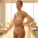 A stunning woman posing confidently in a sun-drenched, high-end modern living room. She has a strong, feminine presence, highlighted by soft, golden-hour natural lighting that accentuates her toned physique and glowing, tanned skin. Her makeup is glamorous and polished, featuring defined winged eyeliner and glossy lips. She is wearing a delicate champagne-colored silk lace bralette and matching high-waisted briefs, layered with a sheer, flowing robe that adds an elegant, ethereal quality. The background showcases a blurred, minimalist interior design with large floor-to-ceiling windows, creating a luxurious and serene atmosphere.