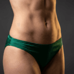 A hyper-realistic, 8k editorial photograph captured with an 85mm macro lens, focusing on the athletic midsection of a woman. The lighting is masterfully executed using a dramatic, high-contrast hard-light setup from the side, which accentuates the defined abdominal muscles and the 'snatched' waistline with sharp, contour-defining shadows. The skin texture is rendered with clinical precision, exhibiting a subtle, natural luminescence and a healthy, hydrated sheen. The subject wears deep emerald-green satin-finish bikini bottoms, the fabric catching the light with a soft, luxurious luster. The composition is a tightly cropped, studio-grade shot set against a minimalist, dark charcoal background, creating a sleek, high-fashion, and sophisticated aesthetic.