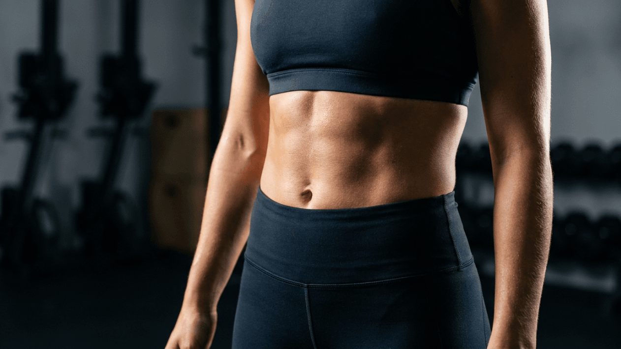 A professional, high-definition editorial shot featuring the torso of a female athlete, captured with an 85mm macro lens to achieve a shallow depth of field and hyper-realistic clarity. The dramatic side-lighting emphasizes her defined, athletic abdominal muscles and core strength with sharp, high-contrast shadows. Her skin exhibits a subtle, healthy luminescence, lightly glinting with a sheen of moisture consistent with post-workout exertion, highlighting the intricate texture and muscle definition. She is dressed in sleek, matte-black high-performance athletic wear, consisting of a sports bra and high-waisted leggings crafted from a technical, compression-fit fabric. The composition focuses intently on the midsection, while the background—a blurred, dimly lit fitness studio with industrial gym equipment—is rendered in deep, muted tones, creating an elite, focused, and powerful atmosphere.