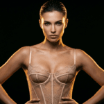 A hyper-realistic 8K editorial portrait featuring a woman with a powerful, confident stance, captured with a professional 85mm prime lens for optimal portrait compression. The lighting design utilizes intense hard-light rim illumination from side angles, creating dramatic shadows that contour her toned physique and defined facial structure. Her skin exhibits a captivating luminescence, enhanced by a subtle, dewy sheen that catches the golden highlights. She wears a meticulously crafted, nude-toned lace bustier, showcasing intricate embroidery and structured boning. The composition is centered against a deep, void-black background, creating a high-contrast noir atmosphere that elevates the subject's striking presence and sophisticated elegance.