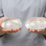 A professional, clinical editorial photograph capturing two breast implants held in the palms of a person wearing a light grey medical scrub top. Shot with an 85mm macro lens, the image maintains a hyper-realistic quality with sharp, 8k resolution. Soft, diffused studio lighting highlights the translucent, gel-like texture and smooth, contoured surfaces of the medical-grade silicone implants. The focus is tightly locked on the objects, emphasizing their structural design and internal details, including the slight variations in shape between the two. The background is a clean, neutral, light-grey backdrop that conveys a sterile, trustworthy, and modern medical environment. The composition is balanced and centered, prioritizing clarity and technical precision to showcase the high-quality materials and craftsmanship of the surgical prosthetics.