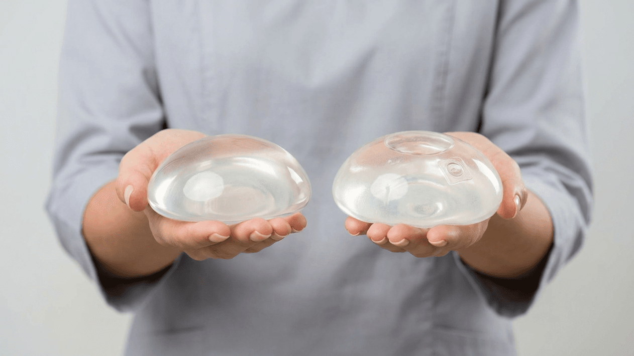 A professional, clinical editorial photograph capturing two breast implants held in the palms of a person wearing a light grey medical scrub top. Shot with an 85mm macro lens, the image maintains a hyper-realistic quality with sharp, 8k resolution. Soft, diffused studio lighting highlights the translucent, gel-like texture and smooth, contoured surfaces of the medical-grade silicone implants. The focus is tightly locked on the objects, emphasizing their structural design and internal details, including the slight variations in shape between the two. The background is a clean, neutral, light-grey backdrop that conveys a sterile, trustworthy, and modern medical environment. The composition is balanced and centered, prioritizing clarity and technical precision to showcase the high-quality materials and craftsmanship of the surgical prosthetics.