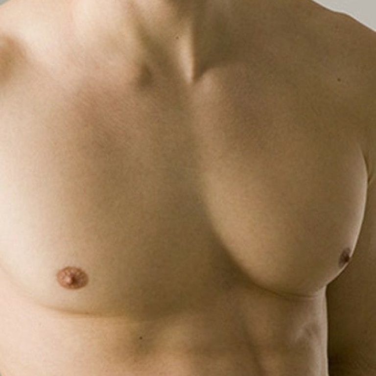 A close-up studio shot of a shirtless man's torso. The lighting is soft and even, highlighting well-defined pectoral muscles and a toned abdomen. The subject exhibits masculine anatomy, with no visible makeup, against a clean, neutral, light grey background. He is not wearing any clothing.