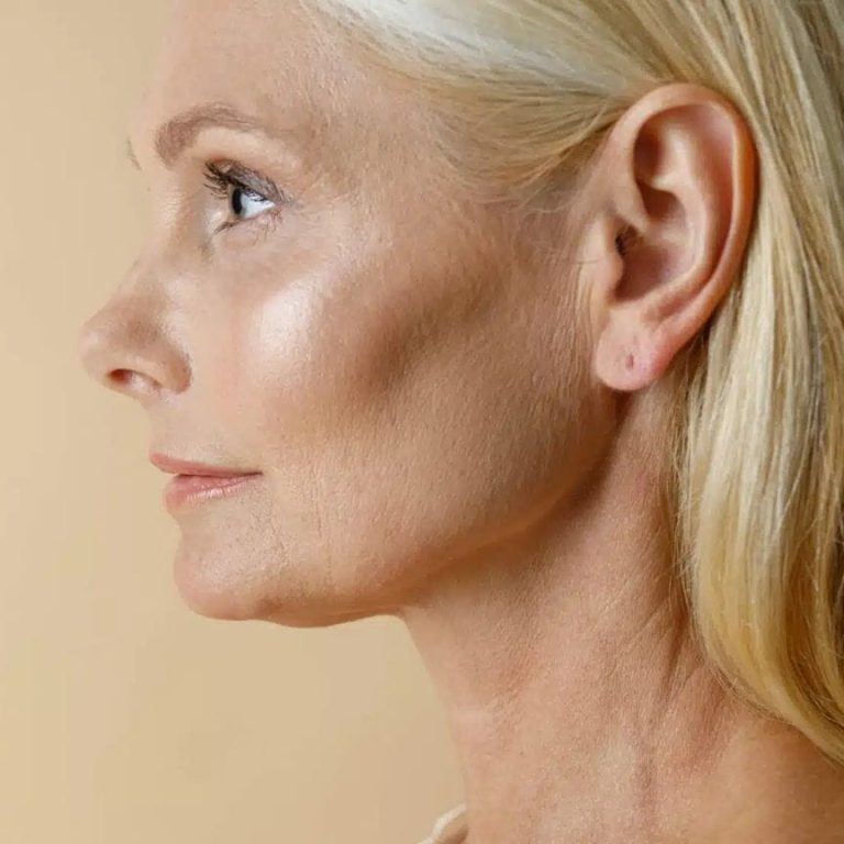 A close-up side profile of an elegant, mature woman with blonde hair, soft natural lighting highlighting her facial features, wearing minimal, fresh-faced makeup that emphasizes a feminine aesthetic, set against a smooth, neutral beige background; her anatomy shows graceful, aged skin texture and sharp bone structure, dressed in a simple, light-colored garment.