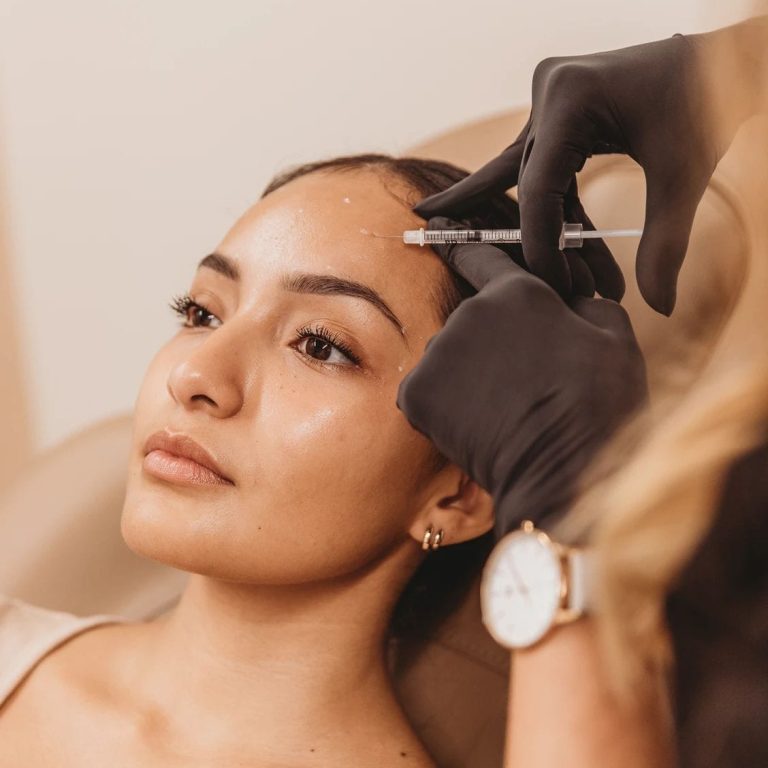 A close-up shot captures a professional aesthetic procedure where a female client with natural, minimal makeup receives a cosmetic injection in her forehead. The lighting is soft and warm, emphasizing the smooth texture of her skin and feminine facial anatomy. The practitioner, wearing black medical gloves, is shown partially in the foreground, creating a contrast with the client's light, neutral-colored top. The background is a clean, neutral beige, providing a professional and calming clinic environment. Focus is on the precision and hygiene of the aesthetic treatment.