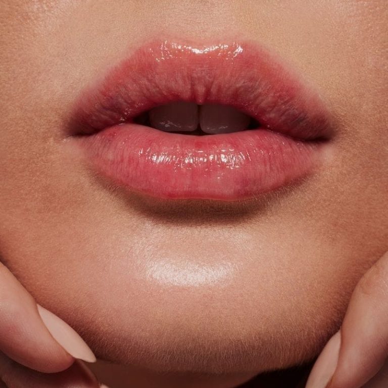 Close-up shot of feminine lips with a glossy, natural-toned finish. The lighting is soft and even, highlighting the skin's texture and a slight shimmer on the chin. The makeup is minimalist and natural, emphasizing the lips' hydrated appearance. The subject is holding their chin with hands featuring nude-colored, manicured almond-shaped nails. No clothing is visible in the tightly framed shot, and the background is a solid, warm neutral tone. The focus is on anatomical lip structure and skin health.