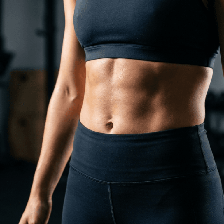 A professional, high-definition editorial shot featuring the torso of a female athlete, captured with an 85mm macro lens to achieve a shallow depth of field and hyper-realistic clarity. The dramatic side-lighting emphasizes her defined, athletic abdominal muscles and core strength with sharp, high-contrast shadows. Her skin exhibits a subtle, healthy luminescence, lightly glinting with a sheen of moisture consistent with post-workout exertion, highlighting the intricate texture and muscle definition. She is dressed in sleek, matte-black high-performance athletic wear, consisting of a sports bra and high-waisted leggings crafted from a technical, compression-fit fabric. The composition focuses intently on the midsection, while the background—a blurred, dimly lit fitness studio with industrial gym equipment—is rendered in deep, muted tones, creating an elite, focused, and powerful atmosphere.