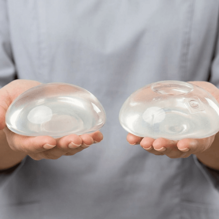 A professional, clinical editorial photograph capturing two breast implants held in the palms of a person wearing a light grey medical scrub top. Shot with an 85mm macro lens, the image maintains a hyper-realistic quality with sharp, 8k resolution. Soft, diffused studio lighting highlights the translucent, gel-like texture and smooth, contoured surfaces of the medical-grade silicone implants. The focus is tightly locked on the objects, emphasizing their structural design and internal details, including the slight variations in shape between the two. The background is a clean, neutral, light-grey backdrop that conveys a sterile, trustworthy, and modern medical environment. The composition is balanced and centered, prioritizing clarity and technical precision to showcase the high-quality materials and craftsmanship of the surgical prosthetics.
